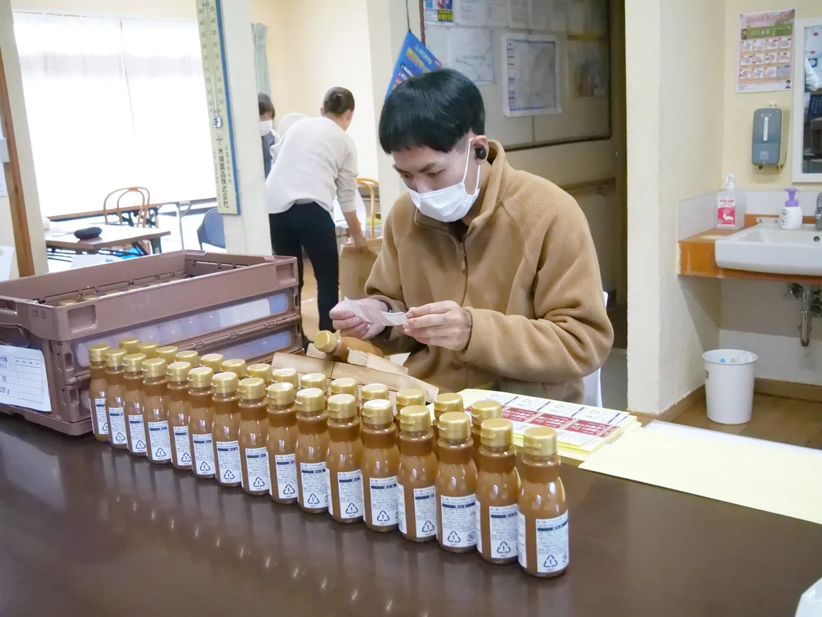瓶商品が並ぶ作業台でラベルを手に取り、貼る準備をする人物