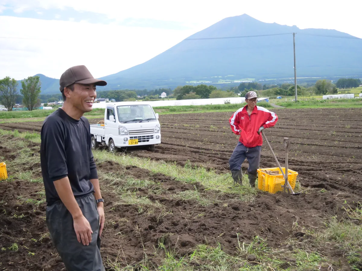 岩手山を背景に、農具や収穫かごをそばに置いてじゃがいも畑で休憩する2人