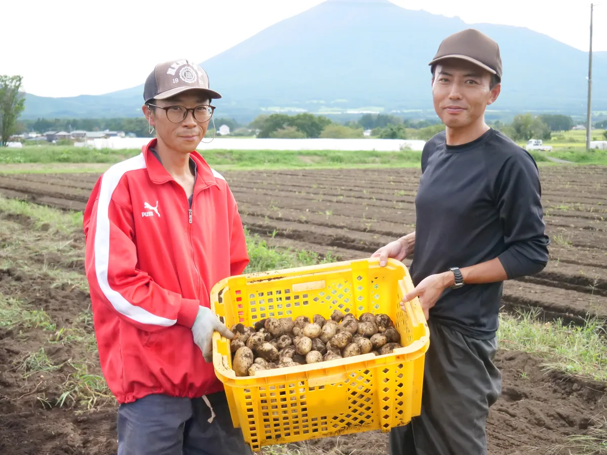山とじゃがいも畑を背景に、じゃがいもが入った黄色いかごを持つ2人